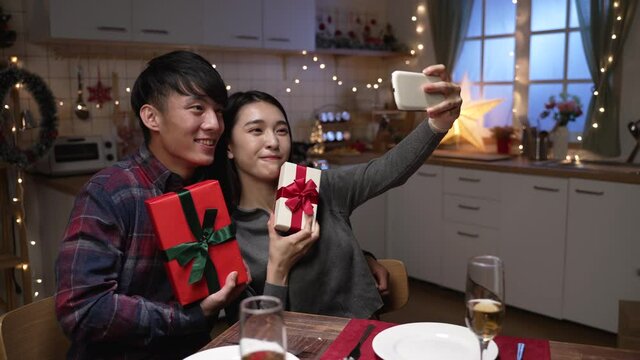 Happy young couple doing selfie using smart phone holding gift boxes and smiling on sweet kitchen interior. lady taking self photo with boyfriend on date night by cellphone