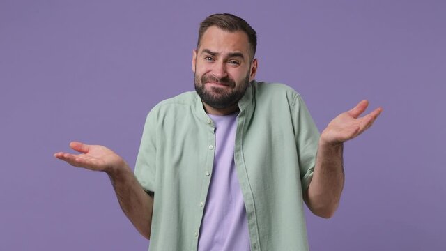 Fun confused shy shamed pensive young bearded man 20s years old wear mint shirt look camera spreading hands say oops ouch oh omg i am so sorry isolated on plain light purple background studio portrait