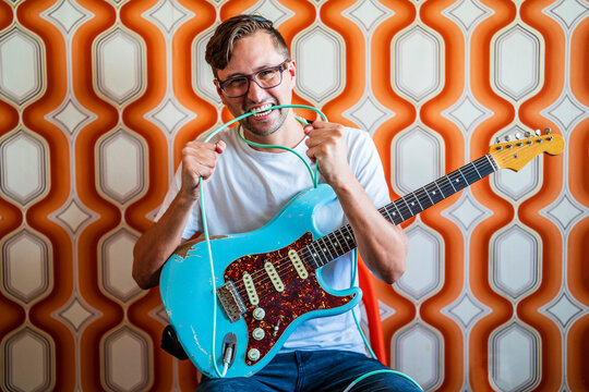 Young male guitarist biting guitar cable in front of wall