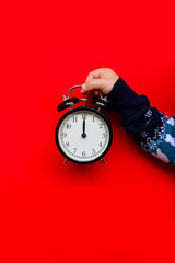 A small child in a Christmas sweater is holding a black vintage alarm clock on a red background. New Year's card. It's time for a holiday and gifts. Midnight on the clock
