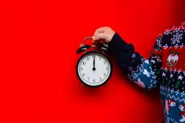 A small child in a Christmas sweater is holding a black vintage alarm clock on a red background. New Year's card. It's time for a holiday and gifts. Midnight on the clock
