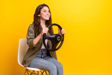 Portrait of attractive cheerful girl sitting on chair holding in hands steering wheel copy space isolated over vibrant yellow color background