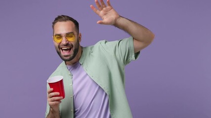 Smiling cheerful happy attractive young bearded man 20s wears mint shirt glasses plastic cup dancing waving hand greeting having fun relax isolated on plain light purple background studio portrait