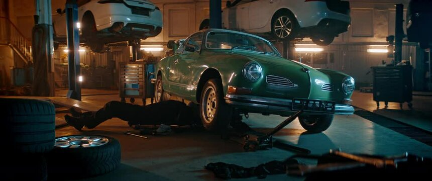 50s Adult Caucasian male mechanic repairing a vintage old car in a workshop, working under car bottom. Shot with 2x anamorphic lens