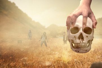 Hand holding a human skull on the field with walking zombies
