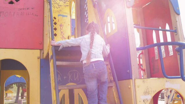 Junior schoolgirl in blue pullover and jeans climbs up wood steps to slide attraction under bright sunlight