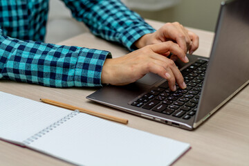 A woman in a plaid shirt is typing on a laptop. There is an empty notebook next to it. The right female hand is in focus. Horizontal view.