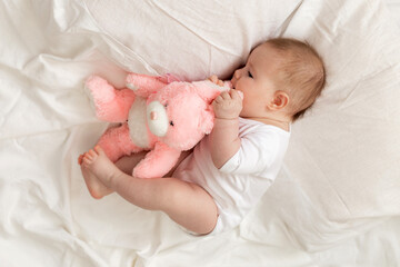 cheerful newborn baby in a white bodysuit lies on a white bed with a plush pink toy rabbit. products for children, toy. concept of a happy childhood and motherhood. child care. space for text.