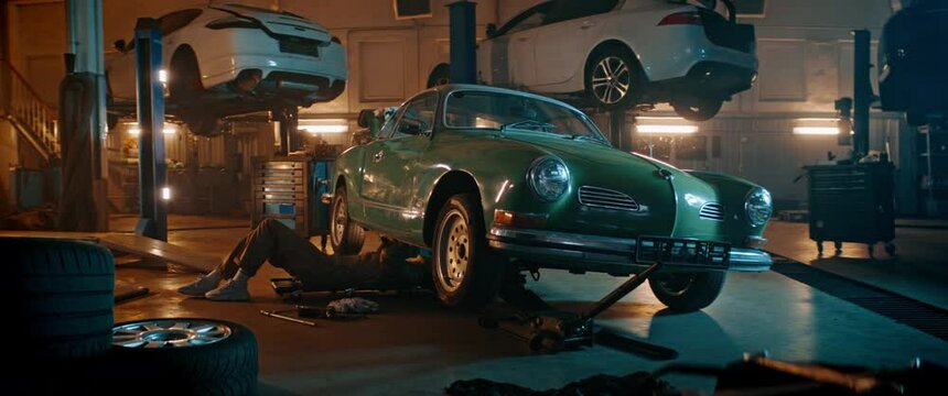 Caucasian female mechanic repairing a vintage old car in a workshop, working under car bottom. Shot with 2x anamorphic lens