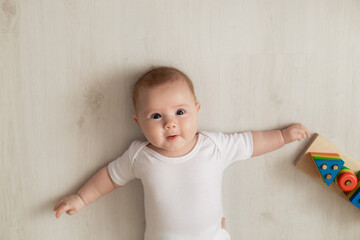 Fototapeta premium cheerful newborn baby in a white bodysuit lies on his back on the floor and plays with educational toys. products for children. concept of a happy childhood and motherhood. child care. space for text