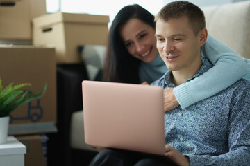 Young married couple communicating on social networks using laptop in apartment with boxes