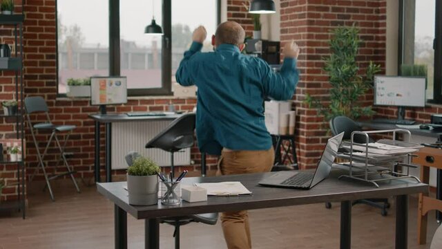 Joyful man celebrating achievement with dance moves and feeling happy about successful win. Employee with face mask dancing after reading good news on laptop and having satisfaction