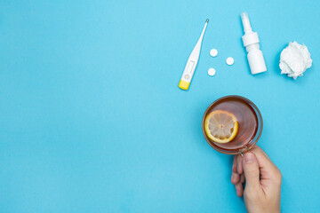 Man holding a cup of tea with lemon. Flu season. Pills, nasal spray, used wipes, and thermometer....