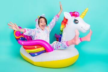 Photo of adorable funky young woman dressed unicorn nightwear smiling riding bloated circle isolated teal color background © deagreez