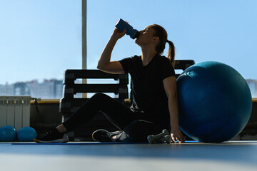 Woman with the Exercise Ball, drinks water from a bottle in the gym, silhouette. Middle-aged  Fitness Woman. Sport and healthy lifestyle concepts.