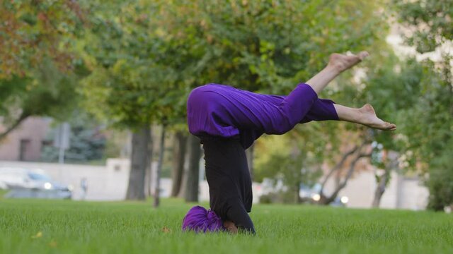 Young strong sporty woman in hijab and purple pants top practicing outdoors, girl trainer doing supported headstand posture, yoga salamba sirshasana asana in park on summer standing on head on grass