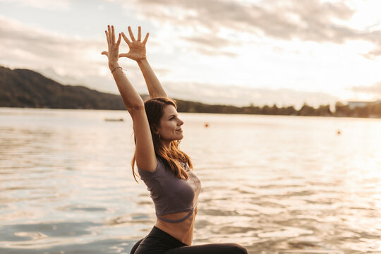 Woman with arms raised doing yoga by lake during sunset