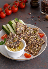 Homemade multigrain seed crackers from ground flax, chia, sesame, sunflower and pumpkin seeds served on a plate with hummus and fresh vegetables. Dark background, selective focus. Ketogenic, vegan