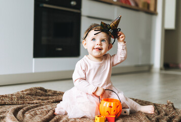 Cute little baby girl in pink dress with the witch's hat and pumpkin lantern sitting on floor in...