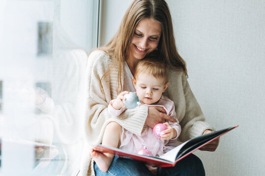 Cute little baby girl in pink dress with her mother young woman reading book in room at home