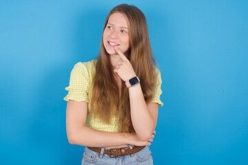 young ukranian girl wearing yellow t-shirt over blue backaground with thoughtful expression, looks to the camera, keeps hand near face, bitting a finger thinks about something pleasant.