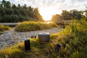 Sunset, dawn on a rocky bank by the river with a camping pot and a thermos, a horizontal frame. Meal on a hike, tourist food. Outdoor activities, summer hikes.