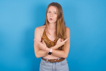 Confused young ukranian girl wearing tank top over blue backaground chooses between two ways, points at both sides with crossed hands, feels doubt. Need your advice.