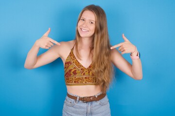 Pick me! Confident, self-assured and charismatic young ukranian girl wearing tank top over blue backaground promoting oneself as wanting role smiling broadly and pointing at body.