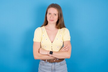 Self confident serious calm young ukranian girl wearing yellow t-shirt over blue backaground stands with arms folded. Shows professional vibe stands in assertive pose.