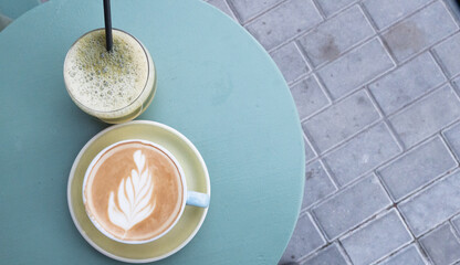 A cup of cappuccino with latte art and and a cocktail with a straw on blue background. Top view with copy spcace