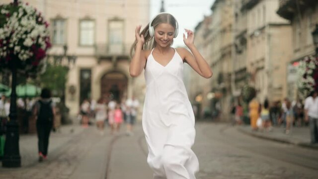 Slow motion of happy young blonde woman in wireless headphones dancing singing outdoors in city street having fun alone. Young adult happy woman in a white dress dancing in downtown very emotional