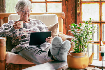 Old senior woman sitting at home on armchair using digital tablet wearing a warm sweater and...