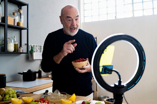 Senior man with fruit bowl vlogging through smart home in kitchen