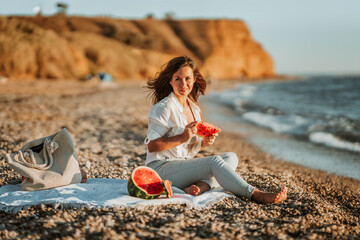 Fototapeta na wymiar A young woman in a white shirt and jeans is sitting on the beach and eating a watermelon. The concept of a quiet summer evening and a picnic with fruit