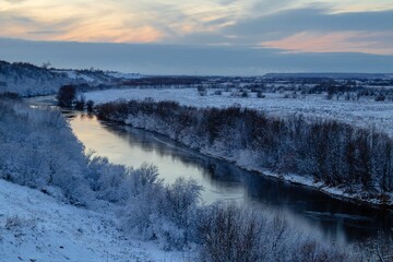 A beautiful winter view from the hill to the river with frosty trees growing along the banks. Winter landscape at sunset