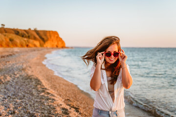 Fototapeta na wymiar Portrait of a happy young woman in a white shirt and red sunglasses walking on the beach at sunset by the sea. The concept of a summer vacation