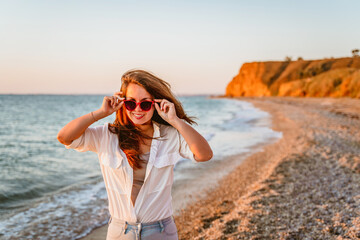 Portrait of a happy young woman in a white shirt and red sunglasses walking on the beach at sunset by the sea. The concept of a summer vacation