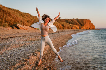 A happy young woman in a white shirt and jeans jumps with her hands up on the beach at sunset. The...