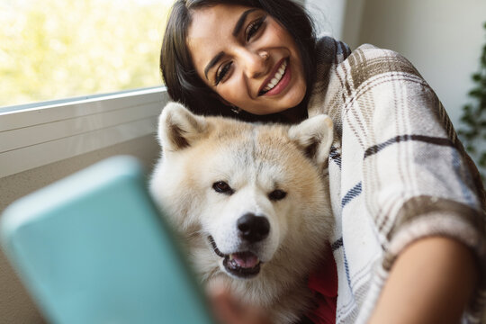Smiling woman with Akita dog taking selfie through mobile phone at home - Powered by Adobe