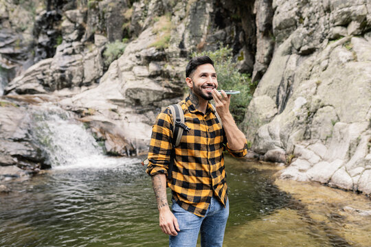 Smiling mid adult man talking on smart phone through speaker by pond