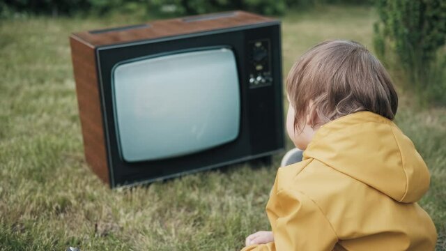 Small child is sitting on grass and watching an old retro TV. TV is strobing interference. Concept of post apocalypse, horror, surrealism and mysticism. Children dependence on TV, gadgets and devices