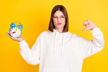 Portrait of attractive moody trendy girl holding clock showing thumb down disapprove isolated over bright yellow color background