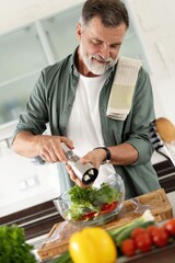 Mature man pouring salt over healthy salad in kitchen for delicious meal. Cooking preparing healthy organic food