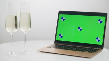 Close-up chromakey laptop with young female hand clinking champagne leaving two glasses on table. Concept of distant holidays celebration with green screen device. Mockup Christmas social distancing