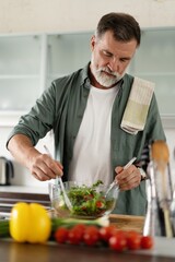 Happy mature man mixing a fresh vegetable salad standing in the kitchen at home.