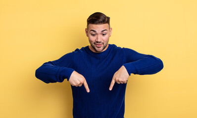 young handsome man with open mouth pointing downwards with both hands, looking shocked, amazed and...