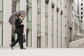 White senior man walking under umbrella at city street