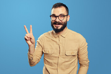 Excited bearded male smiling while showing V sign or victory gesture, wearing eyeglasses and beige...