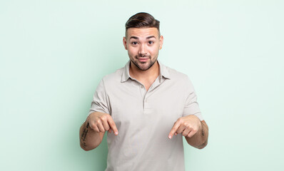 young handsome man with open mouth pointing downwards with both hands, looking shocked, amazed and...