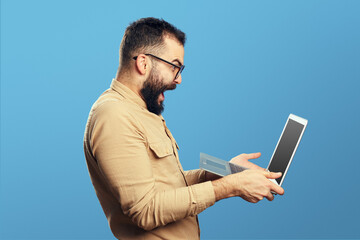 Young bearded male screaming in triumph holding laptop happy with admission to university, isolated over against blue background